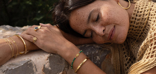 Naturally beautiful mixed race model draped over a rock adorned in Gold bracelets, rings, and earrings from Rachel Entwistle Jewellery Rachel Entwistle